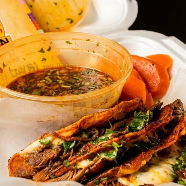 Three cheesy birria tacos with cilantro, a side of consommé, and pickled carrots in a takeout container.