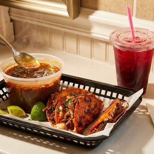 Hand spooning steaming birria consommé, with birria tacos, lime, and a red drink in a basket.